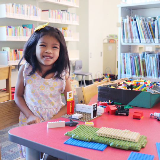 A young child with brown shoulder length hair stands at a table covered in LEGO at McLean Branch. The child is smiling and wearing a floral patterned dress.