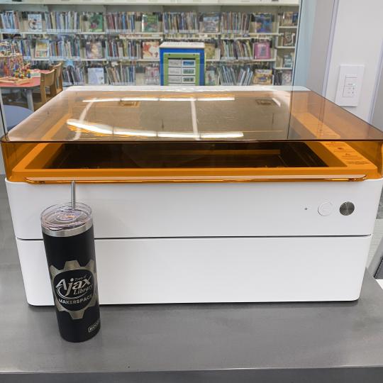 Library's laser cutter, a glowforge, with a tumbler in front of it that has been etched on the laser cutter with the Library's logo.